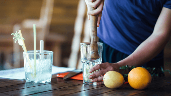 Bartender muddling fruit in glass on wood table with fruit nearby