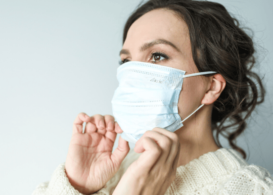 Woman on blank background wearing a white sweater pulling up her face mask