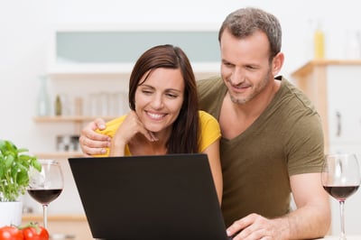 Attractive young couple using a laptop in the kitchen as they enjoy a glass of wine and orange juice together smiling as they read information on the screen